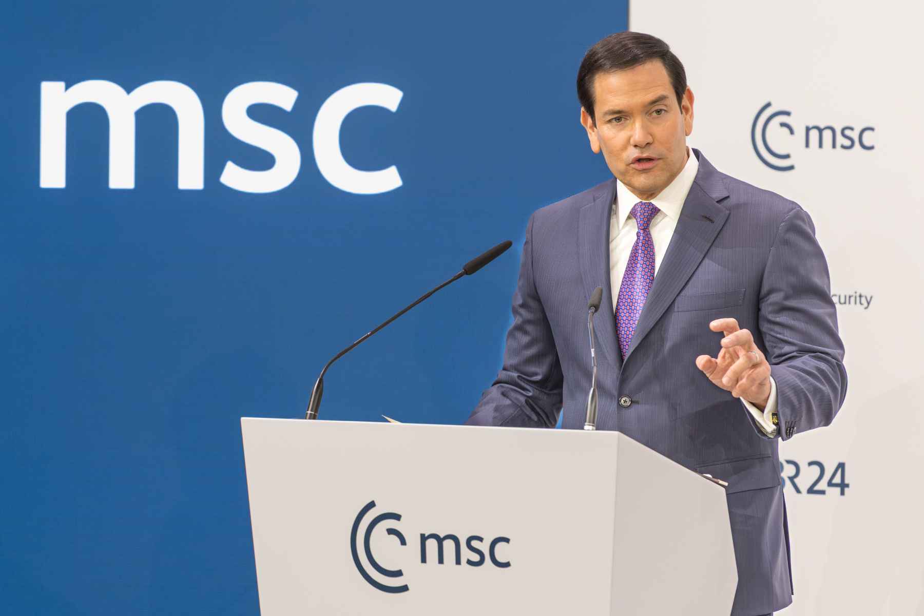 Der Außenminister der Vereinigten Staaten, Marco Rubio, bei einer Rede an einem hellen Stehpult. Darauf und auf einer blauen Wand im Hintergrund die Buchstaben 